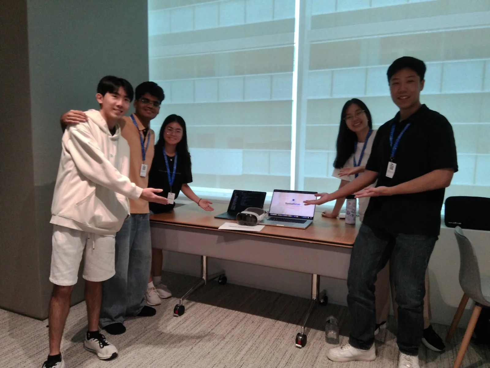 The five-person team presenting SpatialRehab at the Apple Developer Center in Singapore, standing either side of a table holding an Apple Vision Pro and two laptops, one showing the project site.