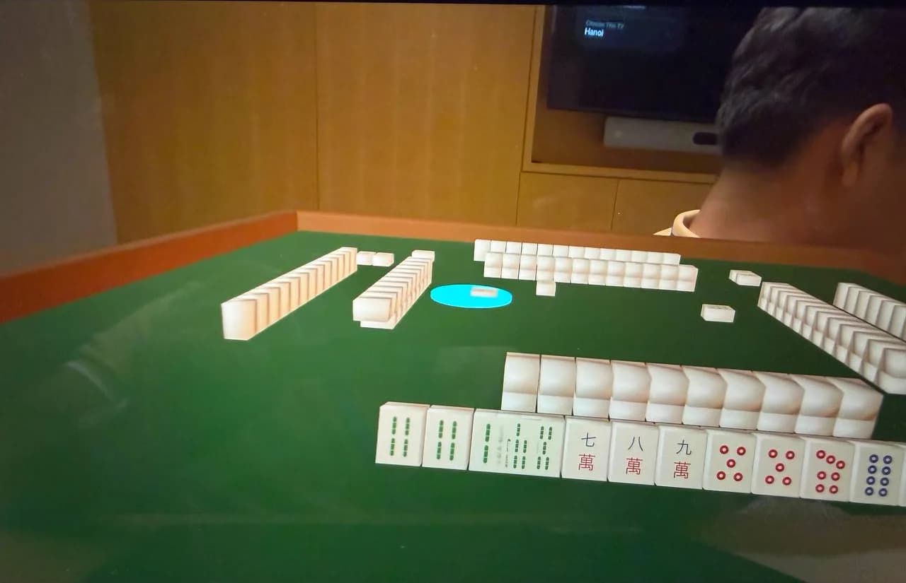 A four-seat Singapore mahjong table rendered in a living room, the player's fourteen tiles laid out in front of them and three computer opponents around the table mid-hand.